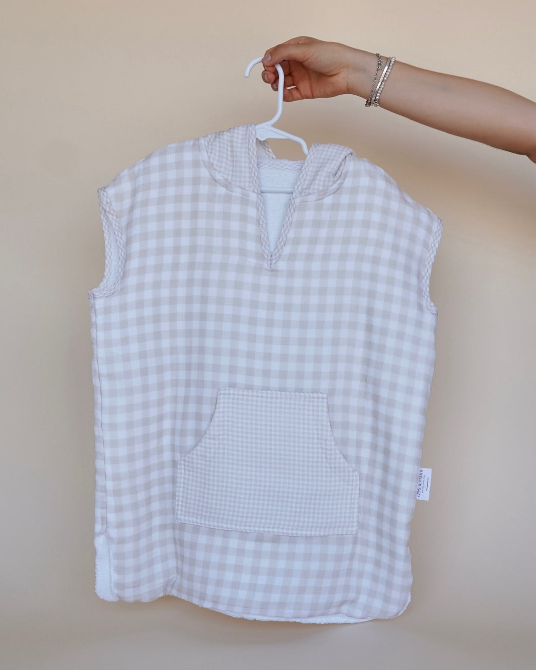 Poncho de toalla y muselina en gancho sostenido por una mano en beige squares