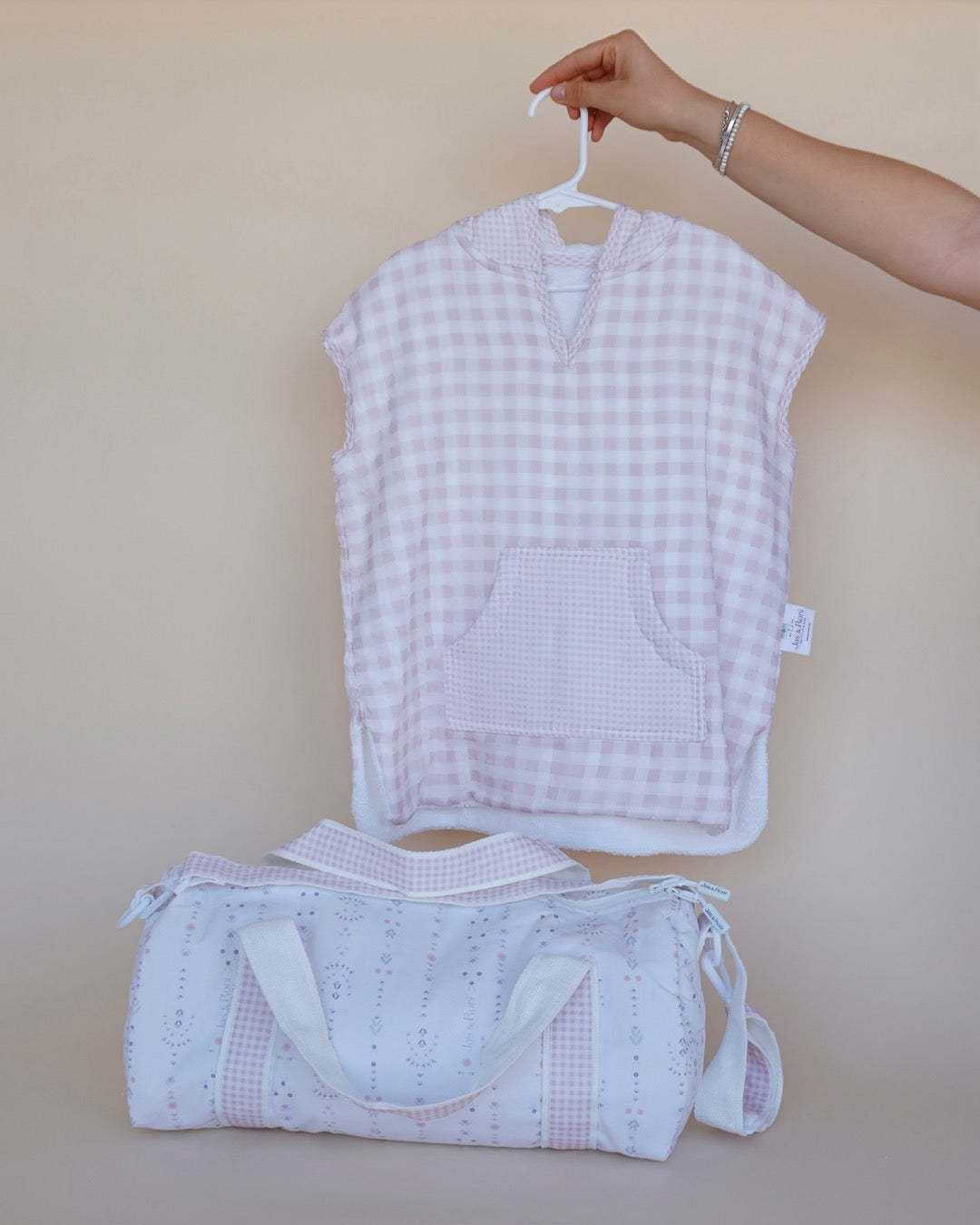 Poncho de toalla y muselina en gancho sostenido por una mano estampado en cuadros rosas y pañalera en tela blossom